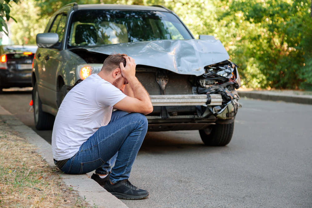 A man stands by his damaged car after an accident, holding his head in frustration and regret.