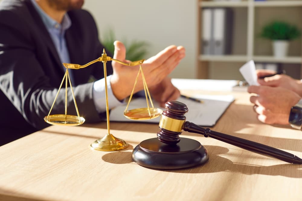 Close-up photo of a male lawyer in his office, discussing case details with a male client at the desk. The scene includes a judge's gavel and scales of justice in the background.