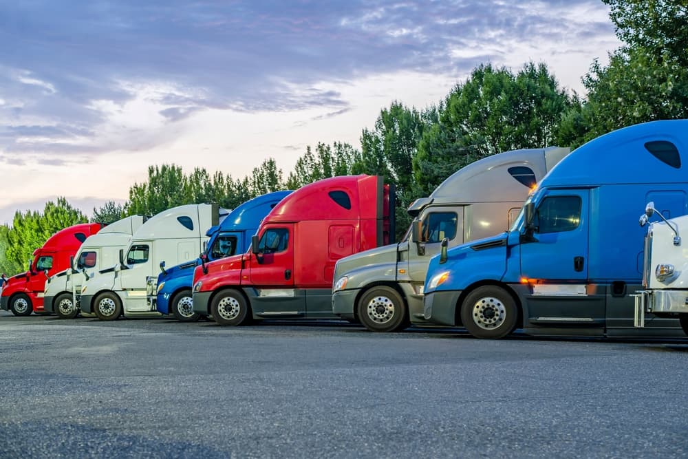 A row of industrial carrier big rig semi-trucks with trailers lined up at a truck stop parking lot, headlights on, resting during twilight hours.