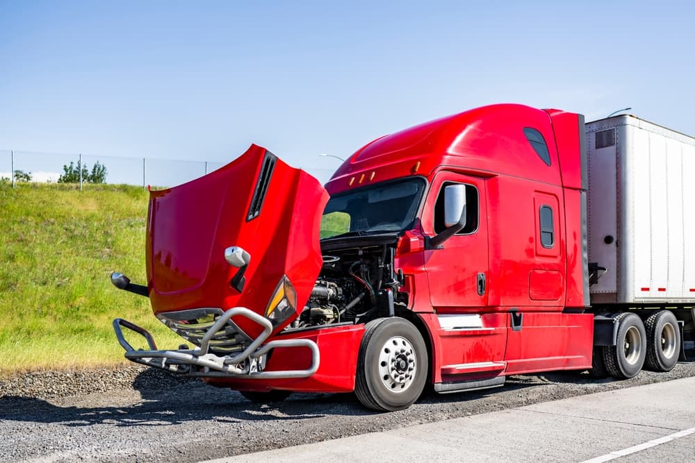 A red broken-down big rig semi-truck with its hood open and attached dry van trailer, parked on the road shoulder, awaiting a tow truck or mobile roadside assistance.